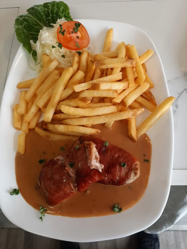 Eine weiße, tiefe Schale mit Gulaschsuppe, die Fleischstücke, Kartoffelwürfel und frische Petersilie enthält. Im Vordergrund auf einem flachen Teller liegen eine goldbraun gebackene Semmel und ein silberner Löffel auf einem hellen Tisch.