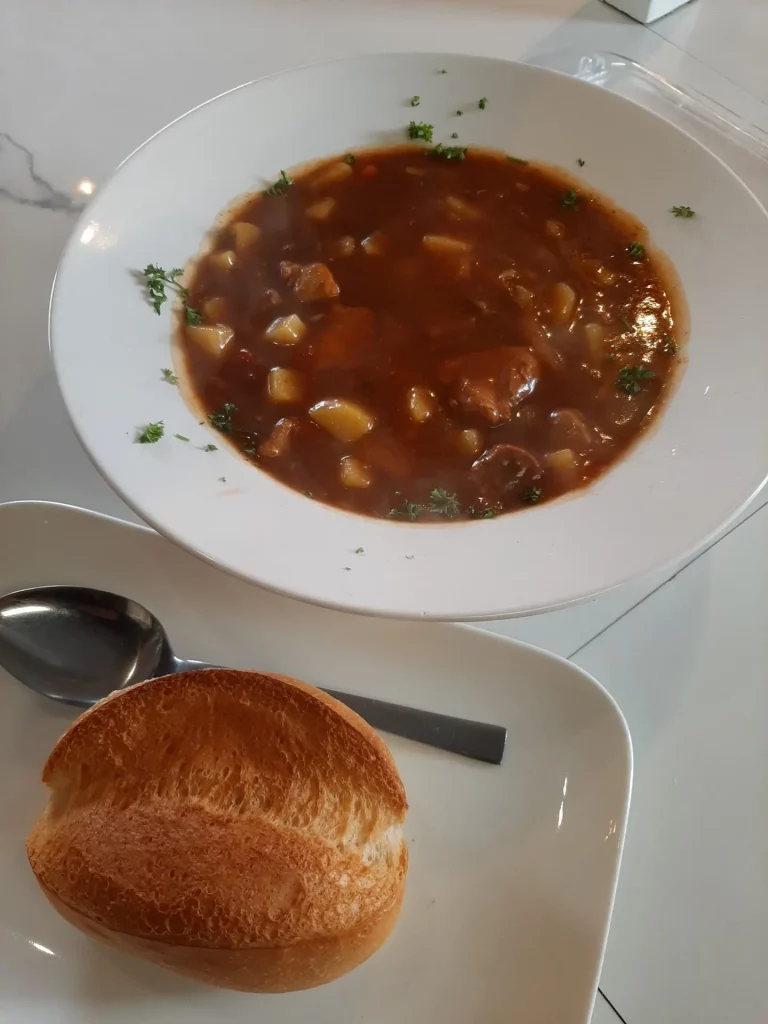 Eine weiße, tiefe Schale mit Gulaschsuppe, die Fleischstücke, Kartoffelwürfel und frische Petersilie enthält. Im Vordergrund auf einem flachen Teller liegen eine goldbraun gebackene Semmel und ein silberner Löffel auf einem hellen Tisch.