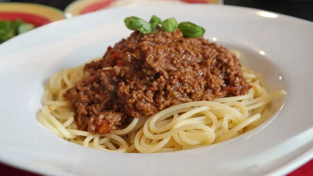 Eine Nahaufnahme einer Portion Spaghetti, die großzügig mit einer dunklen, sämigen Hackfleisch-Tomatensauce (Bolognese) bedeckt ist. Die Pasta ist spiralförmig auf einem tiefen, weißen Teller angerichtet. Obenauf sind fein geriebener Parmesan und ein frisches Basilikumblatt als Garnitur zu erkennen. Der Hintergrund ist leicht unscharf und zeigt einen rustikalen Holztisch und eine Gabel.