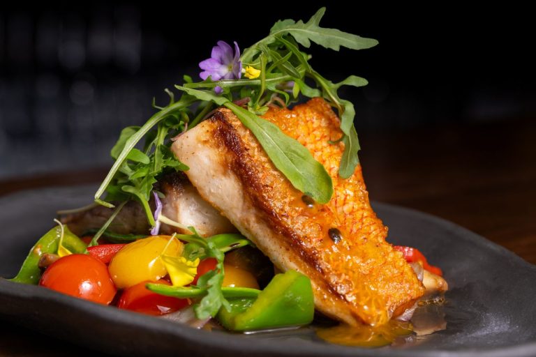 Mittagstisch Freitag, 02.10.2026 4 Delicious grilled fish topped with fresh vegetables and arugula on a dark plate.